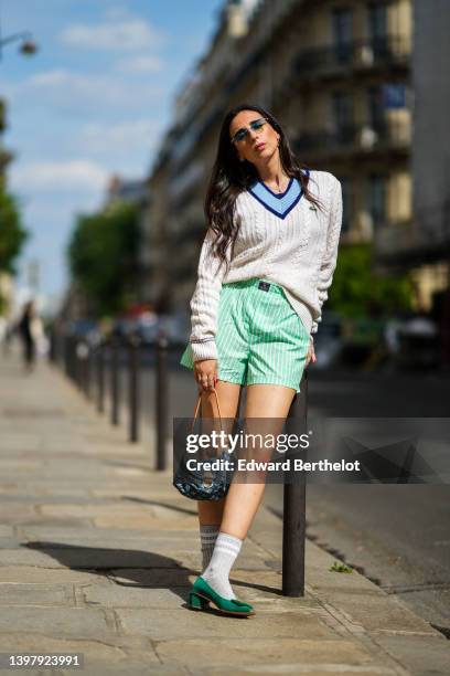 Gabriella Berdugo wears blue sunglasses, gold pendant earrings, a gold chain pendant necklace, a navy blue t-shirt, a white latte braided wool...