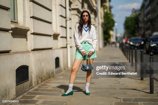 Gabriella Berdugo wears blue sunglasses, gold pendant earrings, a gold chain pendant necklace, a navy blue t-shirt, a white latte braided wool...