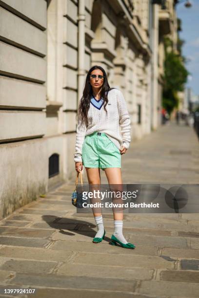 Gabriella Berdugo wears blue sunglasses, gold pendant earrings, a gold chain pendant necklace, a navy blue t-shirt, a white latte braided wool...