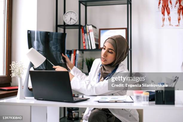 serious asian muslim woman doctor is wearing a lab coat, hijab and a stethoscope discussing on video call over laptop while sitting at clinic. - telemedicine choicepix stock pictures, royalty-free photos & images