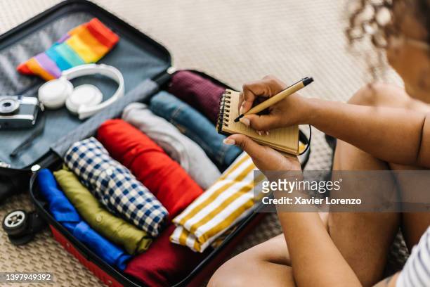 mixed race young adult woman writing list of accessories and travel stuff before going on summer holidays - koffer stockfoto's en -beelden