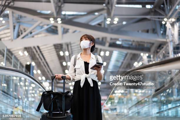 asian young woman wearing a face mask using a phone while standing on the escalator in the airport terminal - thailand covid stock pictures, royalty-free photos & images