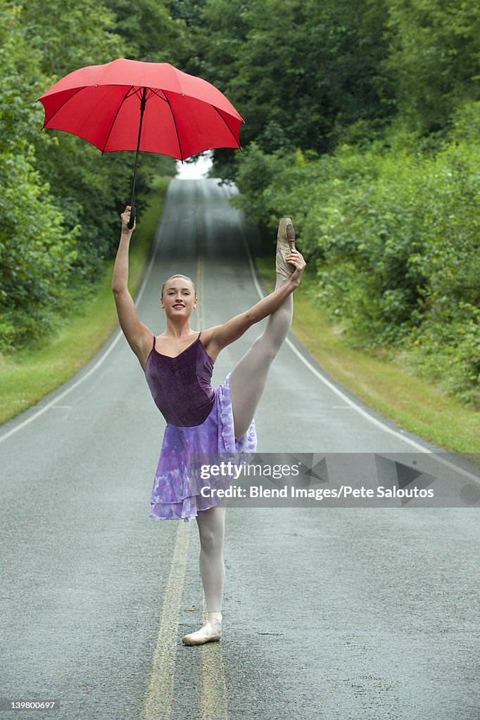 Caucasian ballerina dancing on remote road with umbrella
