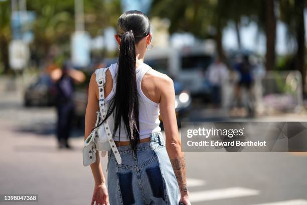 Alexandra Guerain wears gold earrings, a white halter-neck / cropped tank-top, a white shiny leather nailed / studded Le Cagole shoulder bag from...