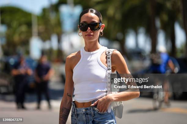 Alexandra Guerain wears black futurist sunglasses from Chanel, gold earrings, a white halter-neck / cropped tank-top, a white shiny leather nailed /...