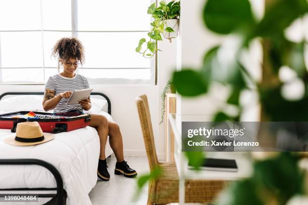 happy young afro american woman using digital tablet sitting on bed packing summer suitcase - booking vacations stock pictures, royalty-free photos & images