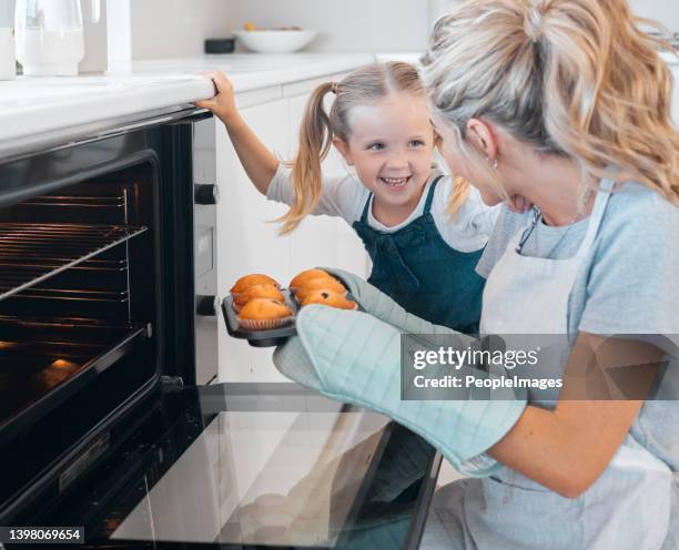Happy mother and daughter removing muffins from the oven. Little girl looking at her parent holding a tray of muffins.Smiling caucasian girl making dessert muffins with her mother.