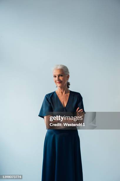 smiling senior woman with arms crossed standing against white background - vestido blanco fotografías e imágenes de stock