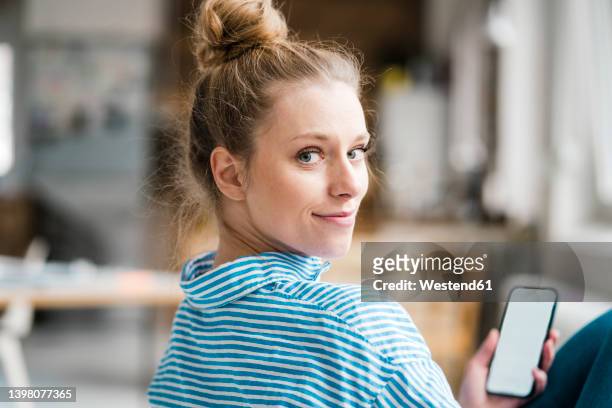smiling freelancer looking over shoulder holding smart phone - mirar por encima del hombro mujer fotografías e imágenes de stock