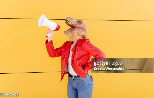 young woman wearing dinosaur mask talking through megaphone in front of yellow wall - dinosaur stock pictures, royalty-free photos & images