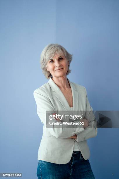 smiling senior businesswoman with arms crossed standing against blue background - encuadre de tres cuartos fotografías e imágenes de stock