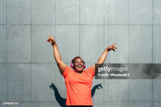 cheerful woman with arms raised enjoying music in front of wall on sunny day - mature women dancing stock pictures, royalty-free photos & images