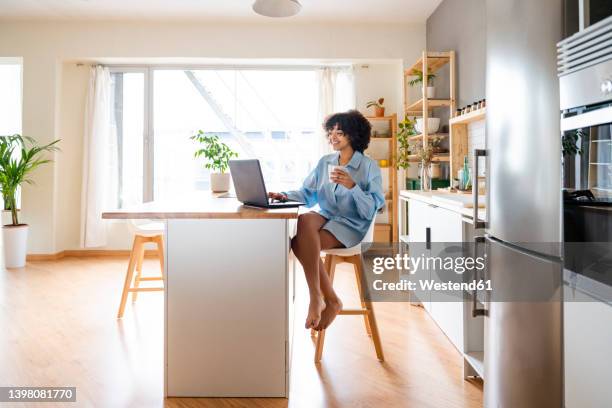 happy woman using laptop sitting on chair at kitchen island - kitchen island stock pictures, royalty-free photos & images