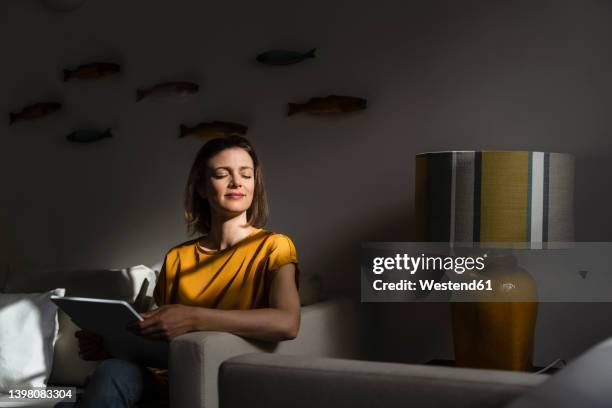 smiling woman with tablet pc sitting on sofa in living room - living room dark stock-fotos und bilder