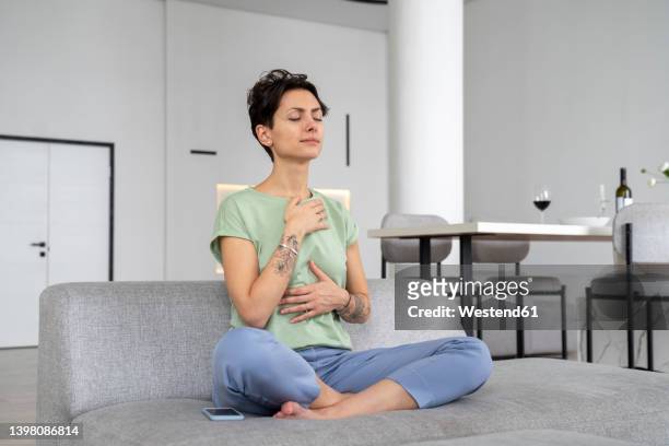woman with eyes closed meditating on sofa at home - atemübung stock-fotos und bilder