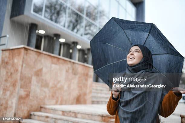 cute muslim female with hijab battling rain with umbrella while taking walk - happy muslim stock pictures, royalty-free photos & images