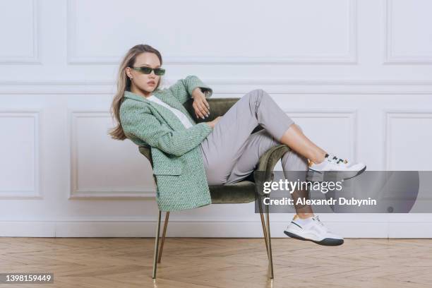 fashion model sitting on chair looking at camera. young blonde woman posing in new collection of fashionable clothes studio shot - beauty cosmetic luxury studio background stock-fotos und bilder