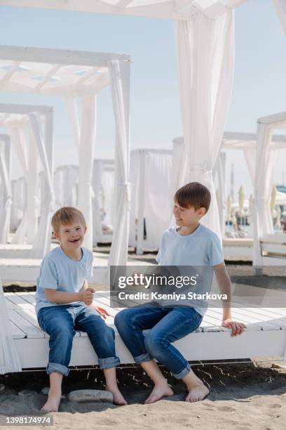 little european fair-haired boy with down syndrome plays with his older brother sitting on a sun lounger on a sandy beach in a blue t-shirt and jeans - fair haired boy stock-fotos und bilder