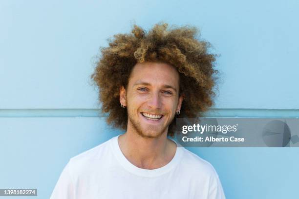 portrait of a young man with afro and blond hair smiling with a blue wall in the background - blonde man stockfoto's en -beelden