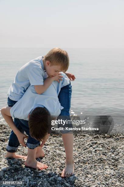 a small european fair-haired boy with down syndrome and his older brother are standing by the sea helping his brother to roll up his jeans - fair haired boy stock-fotos und bilder