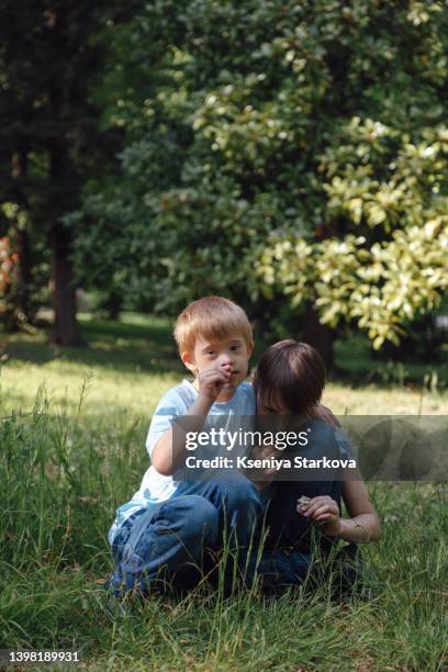 a little european fair-haired boy with down syndrome sits on the grass in the park in a blue t-shirt and jeans holds daisies in his hands together with his older brother look at the camera and smile - fair haired boy stock-fotos und bilder