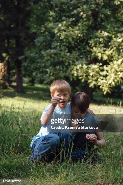 a little european fair-haired boy with down syndrome sits on the grass in the park in a blue t-shirt and jeans holds daisies in his hands together with his older brother look at the camera and smile - fair haired boy stock-fotos und bilder