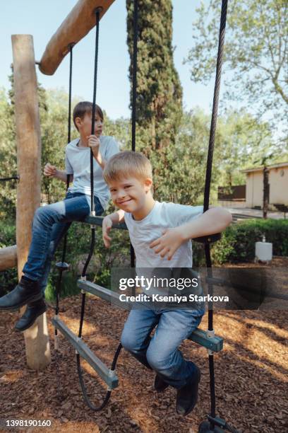 a little european fair-haired boy with down syndrome and his older brother swing on a swing in the park - fair haired boy stock-fotos und bilder