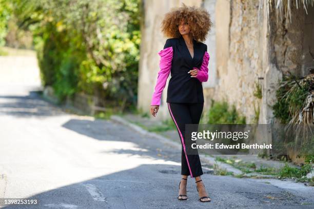 Alicia Aylie wears a black V-neck / pink silk ruffled long sleeves blazer jacket, matching black suit cigarette pants with pink silk straps side,...