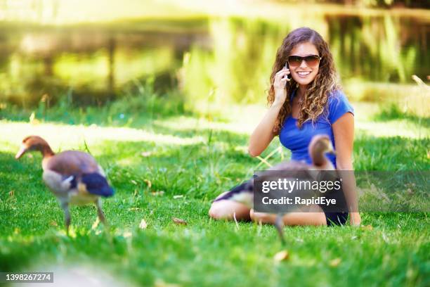 beautiful young woman sits among ducks beside a lake, smiling cheerfully as she talks on her cellphone - ganso do egipto imagens e fotografias de stock