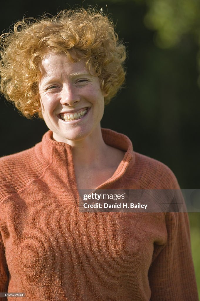 Young red haired woman, 20s-30s, standing in the sunshine, looking at the camera and smiling with a wide happy grin