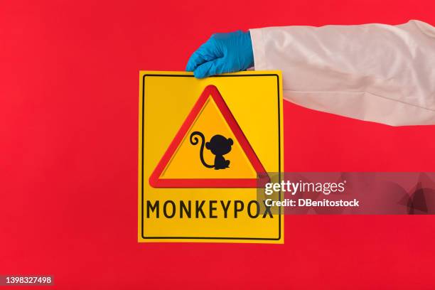 arm of a doctor wearing a gown and latex gloves holding a yellow sign with a symbol with a monkey danger triangle and monkeypox, on a red background. pandemic, virus, epidemic, nigeria and smallpox. - smallpox epidemic stock pictures, royalty-free photos & images