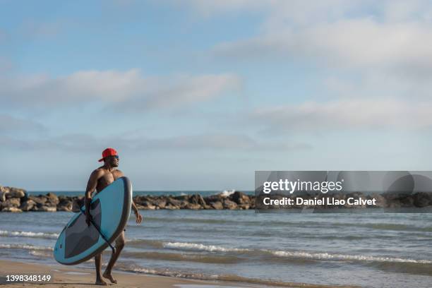 young man holding a paddle board and paddle while walking on the beach. - paddle board men imagens e fotografias de stock