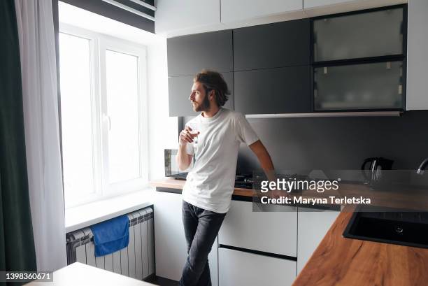 a handsome bearded guy stands and drinks water in the kitchen. modern cuisine. water. bearded guy. casual style. glass of water. - vitamins and minerals imagens e fotografias de stock