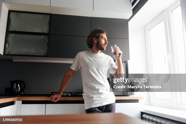 a handsome bearded guy stands and drinks water in the kitchen. modern cuisine. water. bearded guy. casual style. glass of water. - man drinking water fotografías e imágenes de stock