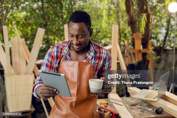 happy young male carpenter working with wood in his workshop in front of laptop during the day. - black man plaid shirt stock pictures, royalty-free photos & images