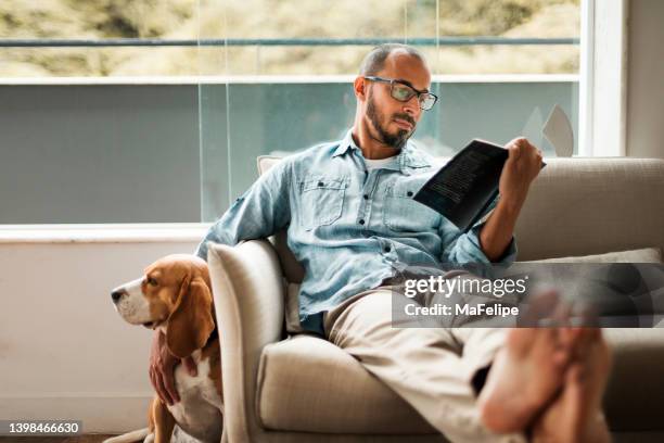 bearded man comfortably sitting on a coach reading a book and holding his dog - middle age man with dog stockfoto's en -beelden