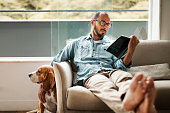 Bearded man comfortably sitting on a coach reading a book and holding his dog