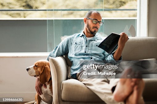 Bearded man comfortably sitting on a coach reading a book and holding his dog Bearded man comfortably sitting on a coach reading a book and holding his dog