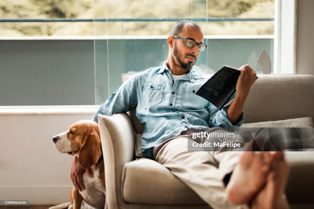 Bearded man comfortably sitting on a coach reading a book and holding his dog