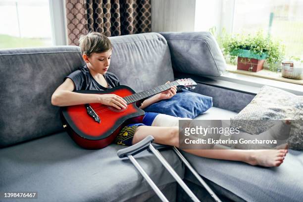 a 12-year-old guy with a broken leg in a cast is sitting on a sofa and playing a red guitar at home. - broken leg stock pictures, royalty-free photos & images