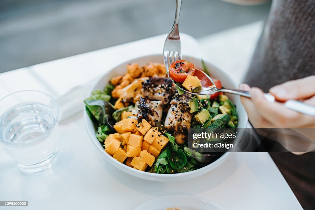 Close up, high angle shot of young woman enjoying multi-coloured healthy fruit, vegetables with grilled chicken salad bowl with balanced nutrition in cafe, with a glass of water by the side. Healthy eating lifestyle. People, food and lifestyle concept