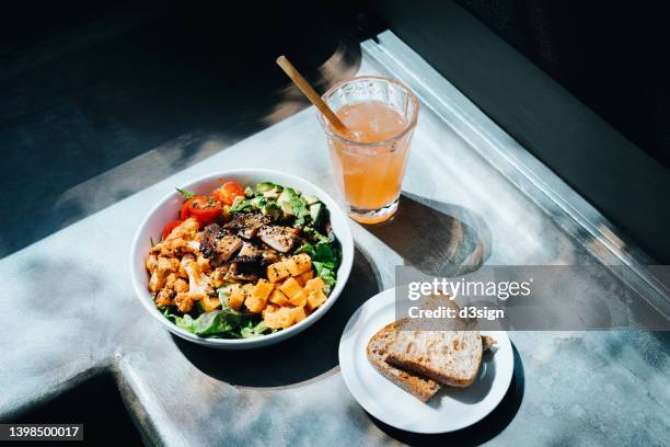 a multi-coloured healthy salad bowl with assorted fresh fruits and vegetables, pumpkin, cauliflower, cherry tomatoes, avocado, lettuce and grilled chicken. with a glass of iced lemonade and sourdough toast by the side on grey background against sunlight - cauliflower bowl stock pictures, royalty-free photos & images