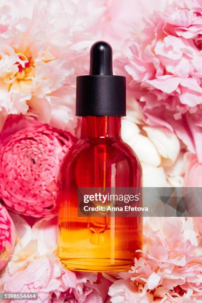 orange glass bottle of face serum with polyglutamic acid is lying on gentle pink peonies and ranunculus. beautiful backdrop for your design. macro photography from above - makeup pile stock pictures, royalty-free photos & images