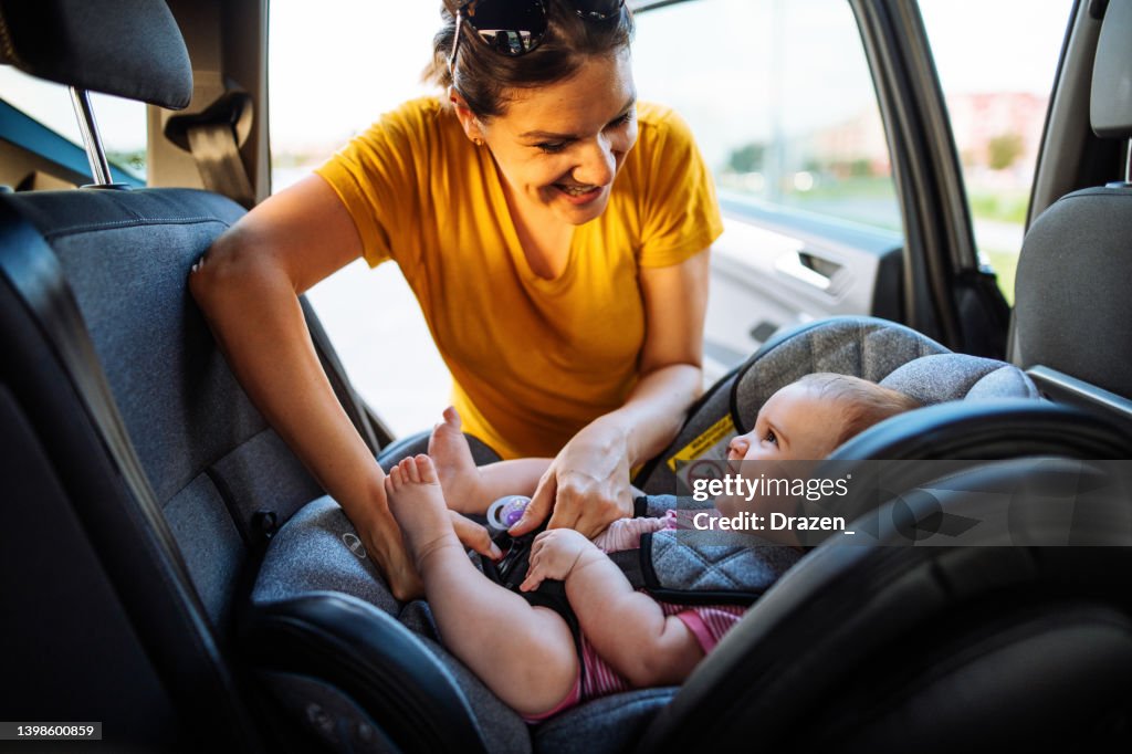 Mère mettant la petite fille dans un siège d’enfant dans la voiture. Famille se préparant pour un road trip en été