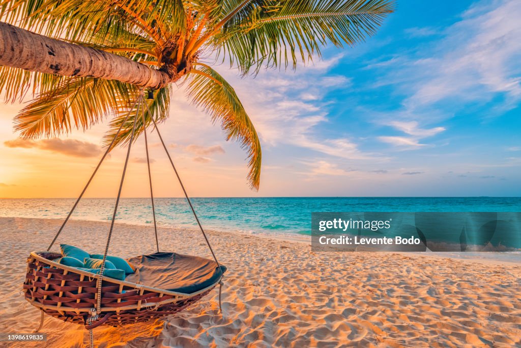 Beautiful tropical sunset beach and sky background as exotic summer landscape with beach swing or hammock and white sand and calm sea beach banner. Paradise island beach vacation or summer holiday destination