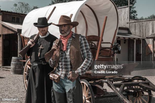 handsome black cowboy with a priest in an authentic wild west setting - handsome cowboy stock pictures, royalty-free photos & images