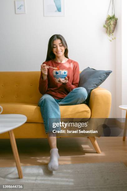 young woman eating a bowl of cereal and fruit while enjoying a relaxing moment on the sofa at home. - eating alone fotografías e imágenes de stock