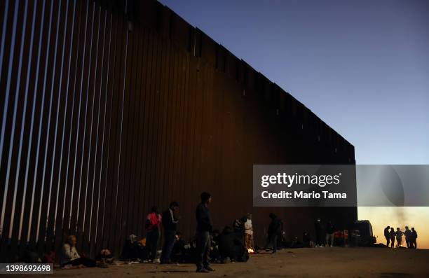 Immigrants from Cuba and Venezuela warm themselves by a fire before sunrise along the U.S.-Mexico border barrier as they await processing by the U.S....