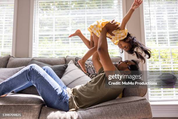 father lifting daughter in the air on couch - asian man barefoot foto e immagini stock