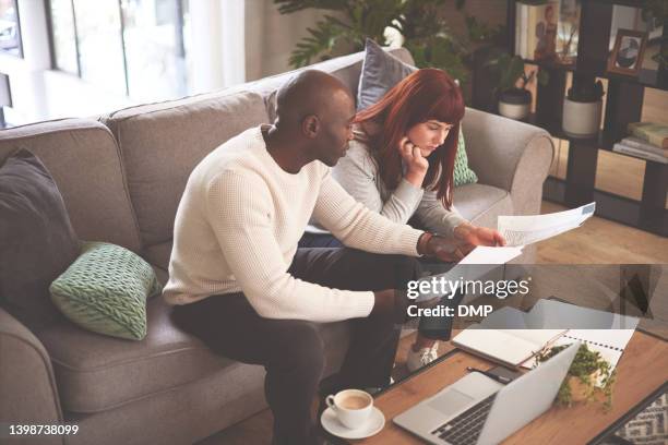 a couple going through paperwork at home and using a laptop. a young man and woman paying bills, planning their finance and looking at paper online with a computer on the sofa - home insurance stock pictures, royalty-free photos & images